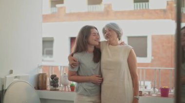 Gümüş saçlı bir anne ve kızının bir balkonda sevgi dolu kucaklaşmalarını gösteren dokunaklı bir sahne. İkisi de bir şehir ortamında hafifçe gülümsüyor, sıcak ve ailevi bir bağ kuruyor.