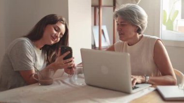 Bir anne ve kızın mutfak masasındaki sıcak ve ilgi çekici sahnesi, akıllı bir telefonla gülümseyen ve etkileşime geçen, dizüstü bilgisayar ve içeceklerle, rahat, ailevi bir atmosferi aktaran