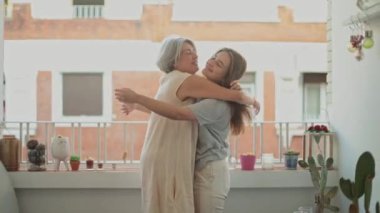 Güneşli bir balkonda kucaklaşan bir anne ve kızın sıcak ve sevgi dolu bir sahnesi.