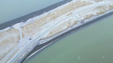 İzlanda 'daki soğuk bataklık arazisinde kıvrılan bir yol arasındaki zıtlığı gösteren keskin bir hava görüntüsü, buzlu maviler ve toprak tonlarının bir karışımını sergiliyor.