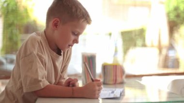 Kafkasyalı bir çocuk, gerçek zamanlı öğrenmeye dalmış, ödevini iyi aydınlatılmış bir odada, elinde bir kalem ve yanında renkli bir kalem kılıfıyla özenle yapıyor..