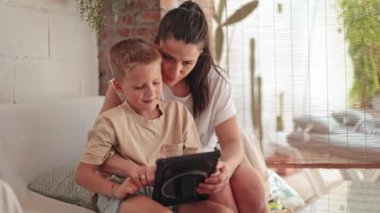 Kafkasyalı bir anne ve küçük oğlu rahat bir ev dekorunun ortasında dijital tabletle meşgul olan rahat, iyi ışıklandırılmış bir balkonda kaliteli zaman geçiriyorlar..