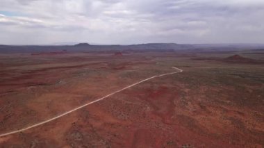 Utah 'taki geniş, kırmızı tonlu çöl manzarası geniş ve bulutlu bir gökyüzünün altında engebeli araziyi keskin bir şekilde kesen uzun ve dolambaçlı bir yola ev sahipliği yapıyor.