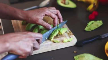 Sağlıklı bir yemek atölyesinde, bir insan diğer meyvelerle çevrili ahşap bir tahtada taze avokadoyu dikkatlice keser.