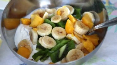 Renkli bir meyve salatası karışımı. İçinde muz, kivi ve mango dilimleri var. Temiz cam kasede kremalı yoğurt katmanlı.