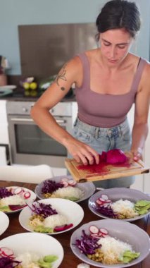 Odaklanmış bir kadın, bir yemek atölyesi sırasında canlı ve sağlıklı yemekler hazırlarken görülüyor. Çeşitli tabaklar üzerinde taze kesilmiş pembe pitaya 'yı dikkatlice düzenliyor.