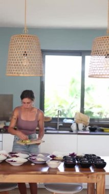 Bir kadın, modern mutfak dekoru ve renkli ürünlerle çevrili sağlıklı bir yemek atölyesi için avokado da dahil olmak üzere taze sebzeleri ve meyveleri dikkatlice düzenler.