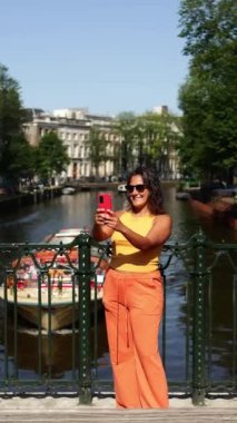 Sarı bluzlu ve turuncu pantolonlu neşeli bir kadın Amsterdam 'da bir kanalda duruyor, kırmızı akıllı telefonuyla selfie çekerken gülümsüyor.