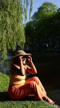 Bir kadın güneşli bir sonbahar gününün tadını çıkarıyor Amsterdam 'da bir kanalın yanında otururken yeşil söğüt ağaçlarıyla çevrili, canlı turuncu bir elbise giymiş şapkasını çıkarıyor.