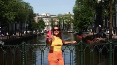 Yaz kıyafetleri içinde neşeli bir kadın, akıllı telefonunu kullanıyor, muhtemelen selfie çekiyor, Amsterdam 'da güzel bir kanalda, arka planda klasik mimarisi ve tekneleri olan bir köprüde.