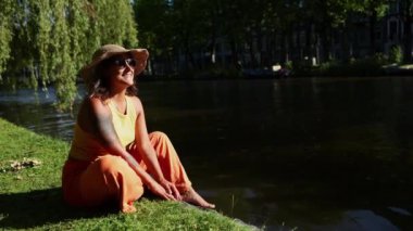Bir kadın, Amsterdam kanalında rahat bir şekilde oturur, sonbahar güneşini emer, hasır şapka takar ve şık güneş gözlüğü takar, günlük kıyafetleriyle etrafındaki canlı sonbahar renkleriyle harmanlanır.