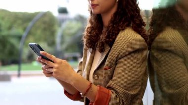 smiling Brazilian with curly hair businesswoman checks her smartphone while standing by a glass wall in an office. She's dressed professionally in a plaid jacket and jeans.