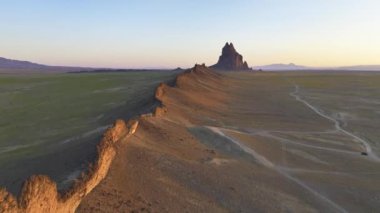 New Mexico 'da ikonik bir kaya oluşumu olan Shiprock' un altın saatinde geniş çöl düzlüklerini ve dolambaçlı bir patikayı gösteren hava aracı tarafından çekilen çarpıcı hava görüntüsü..