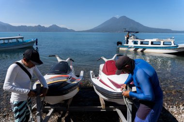 İki İspanyol genç adam Atitlan Gölü kıyısında bir jet-ski hazırlıyor. Bu da su bazlı bir eğlence için hazırlık ve beklenti sürecini gösteriyor.