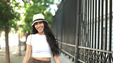 Uzun saçlı, şapkalı ve beyaz bluzlu genç bir bayan ağaçlarla kaplı kaldırımda yürürken parlak bir şekilde gülümsüyor. Güneş ağaçların arasından süzülüp sıcak ve neşeli bir atmosfer yaratıyor..