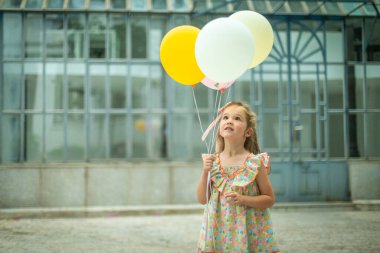 Pembe kurdeleli ve çiçekli elbisesi olan genç bir kız dört renkli balon tutuyor, açık havada dikilirken dik dik dik bakıyor..