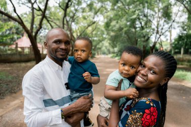 Neşeli bir Afrikalı aile, bir anne ve baba genç oğullarını kucaklıyor, yemyeşil bir parkta gülümsüyorlar. Herkes doğrudan kameraya bakıyor..