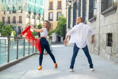Kübalı dansçılar canlı hareketlerini güneşli bir caddede sergiliyorlar. Kadın kırmızı bir eşarp sallarken, erkek dinamik bir poz veriyor, hem neşe hem de enerji saçıyor..