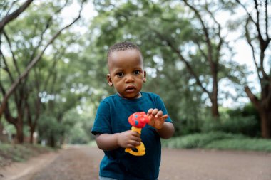 Genç bir Afrikalı çocuk yemyeşil bir parkta kirli bir yolda duruyor, elinde küçük bir oyuncakla kameraya bakıyor..
