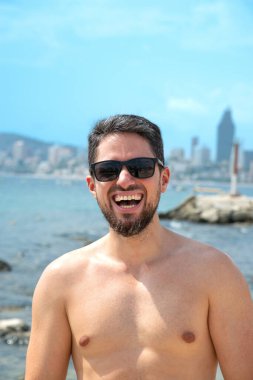 Plajda güneş gözlükleriyle gülümseyen genç İspanyol manken arka planda güzel mavi gökyüzü olan kameraya bakıyor.