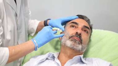 Doktor, erkek hastayı güzellik kliniğinde kozmetik enjeksiyona hazırlıyor. Hastanın yüzü çizgilerle işaretli. Eldiven giyen doktor..