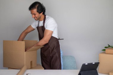Önlük takan bir adam diğer ambalaj malzemeleriyle dolu bir masada büyük bir karton kutu hazırlıyor..