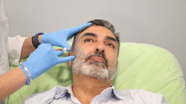 Bir erkek hastanın yüzüne kozmetik enjeksiyon yapan bir doktor, bir güzellik kliniğinde ameliyat için işaretlenmiş bir erkek hastanın yüzüne kozmetik enjeksiyon uyguluyor.