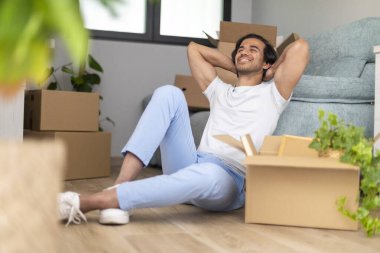 Paketleri açmaya ara veren bir adam, kutular etraftayken rahatça yerde oturuyor..