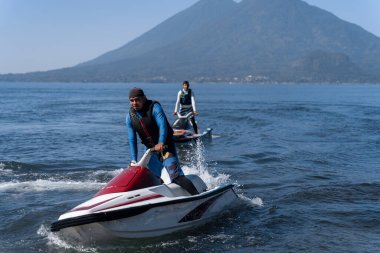 İki İspanyol genç adam, Atitlan Gölü Guatemala 'nın sularında, gölün doğal güzelliğinin manzaralı arka planına karşı eğlence amaçlı su aktivitesine girişerek jet-ski' lerde geziniyorlar.