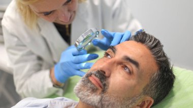 Doktor, bir güzellik kliniğinde bir erkek hastanın yüzünü büyüteçle yakından inceliyor..