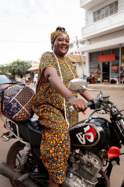 Afrikalı mutlu bir kadının dikey portresi. Motosikletli, renkli bir kadın. Çantasını taşıyor ve Afrikalı bir caddede gülümsüyor.