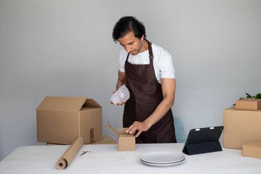 Man wrapping an item in bubble wrap and placing it into a small box, preparing packages on a table with boxes and plates clipart