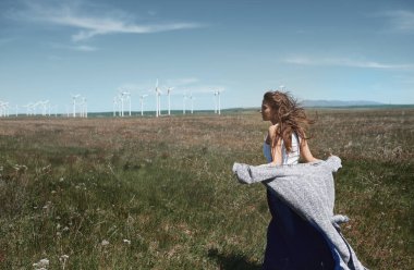 Serene genç kadın, arka planda açık bir alanda duruyor, açık mavi gökyüzünün altında, yenilenebilir enerji ve sükuneti sembolize eden rüzgar türbinleri.