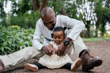 Beyaz gömlekli Afrikalı baba, genç kızıyla yemyeşil bir parkta oynuyor ve ona nasıl alkışlanacağını öğretiyor. İkisi de gülümsüyor, hassas ve neşeli bir anda..
