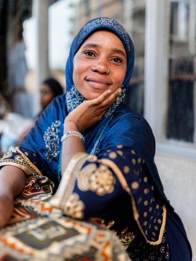 Gülümseyen ve gururlu bir Afrikalı Müslüman kadının dikey portresi açık havada dikiş atölyesinde oturuyor.