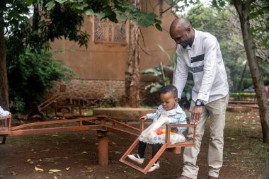 Afrikalı baba genç kızına yemyeşil bir park ortamında tahterevallide kibarca rehberlik ediyor. Kızım, odaklanmış ve aşağıya bakıyor, babasının tetikte bakışları altında neşeli anın tadını çıkarıyor..