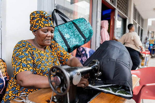 Африканская женщина в традиционной одежде работает шитьем с традиционной машиной в городе, сидя за пределами здания
