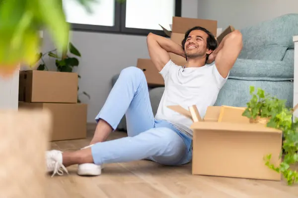Paketleri açmaya ara veren bir adam, kutular etraftayken rahatça yerde oturuyor..