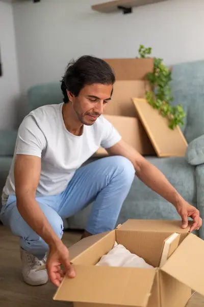 Oturma odasında bavulunu açarken diz çöküp yeni yerini düzenleyen bir adam..