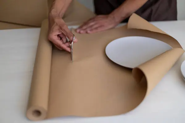Kişi ambalaj tabakları için kahverengi kağıt kesiyor, eller ve malzemelere odaklanıyor.