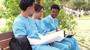 Çeşitli etnik geçmişleri temsil eden üç hemşirelik öğrencisi, yeşillik, dosya ve dijital tabletlerle çevrili bir bankta çalışıp işbirliği yapıyorlar.