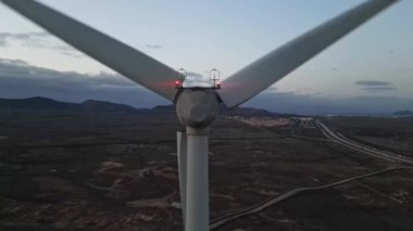 Alacakaranlıkta faaliyet gösteren bir rüzgar türbini görüntülüyor. Etrafı geniş ve uzak bir araziyle çevrili. Yollarla çevrili ve uzakta sönük şehir ışıkları var.