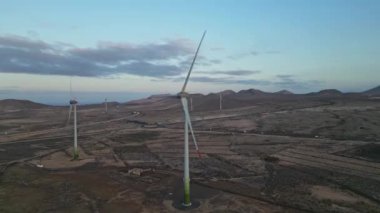 Uçsuz bucaksız bir gökyüzünün altındaki kurak, tepe noktasında çalışan birden fazla rüzgar türbini yakalayan geniş bir çekim, yenilenebilir enerji üretimini vurguluyor.
