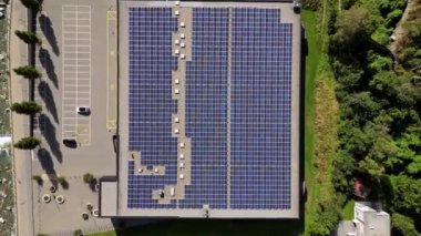 Bereketli yeşil bir bölgenin yanında tam teçhizatlı güneş panelleriyle dolu büyük bir çatı katının havadan çekilmiş görüntüsü, kentsel ortamlarda sürdürülebilir enerji entegrasyonunu gösteriyor