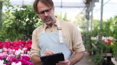 Çizgili önlüklü bir bahçıvan bereketli çiçeklerle dolu bir serada envanteri yönetmek için tablet kullanır. Parlak güneş ışığı çeşitli bitki sırasını aydınlatır.