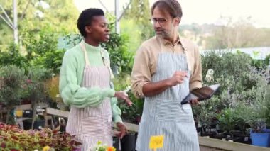 Önlüklü iki bahçe çalışanı bir bitki bakım odasında sohbet ediyorlar. Çeşitli saksı bitkileri ve çiçeklerle çevrili, meşgul ve neşeli görünüyorlar.