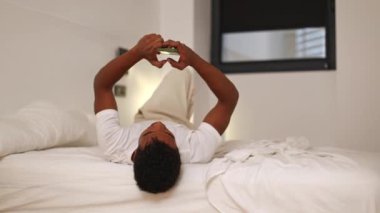 Siyah bir adam bir yatakta rahat bir şekilde uzanır, akıllı telefonuna odaklanır oda hafif ışıklandırmayla minimalisttir, rahatlama ve teknoloji kullanımı için rahat bir ortam yaratır.