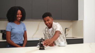 Kadın modern bir mutfakta bir çift olarak rahatlama anını seyrederken, siyah bir adam fincanlara kahve dolduruyor. Sahne, rahat bir sabah rutininin özünü yakalar..