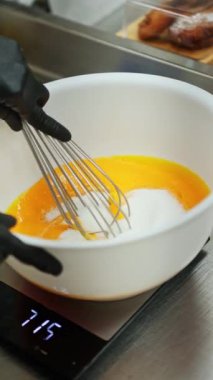 Siyah eldiven giyen biri canlı bir portakal hamuru çırpıyor, muhtemelen şeker ve yumurta sarısı, mutfak tezgahındaki beyaz bir kasede, pişirme ya da pişirme işlemini yakalıyor.