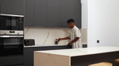 Bir adam koyu dolapları ve mermer tezgahları olan şık, modern bir mutfakta kahve makinesinden fincana kahve dolduruyor, minimalist ve şık bir ortam yayıyor.